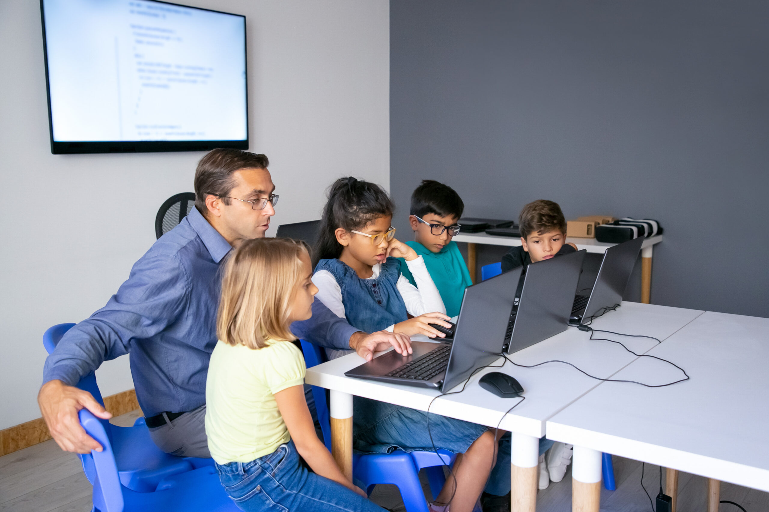 Multiethnic pupils doing task with help of teacher. Concentrated man in glasses sitting with children at table and looking at laptop screen. Childhood, communication and digital education concept